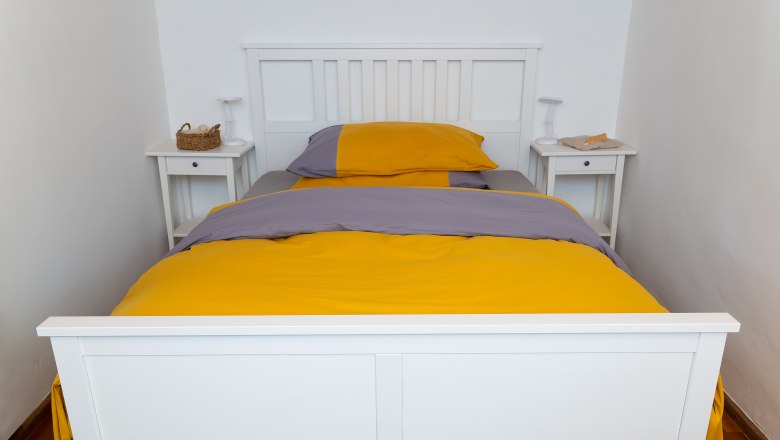 Ein weißes Bett mit gelb-grauer Bettwäsche in einem schlichten Schlafzimmer.