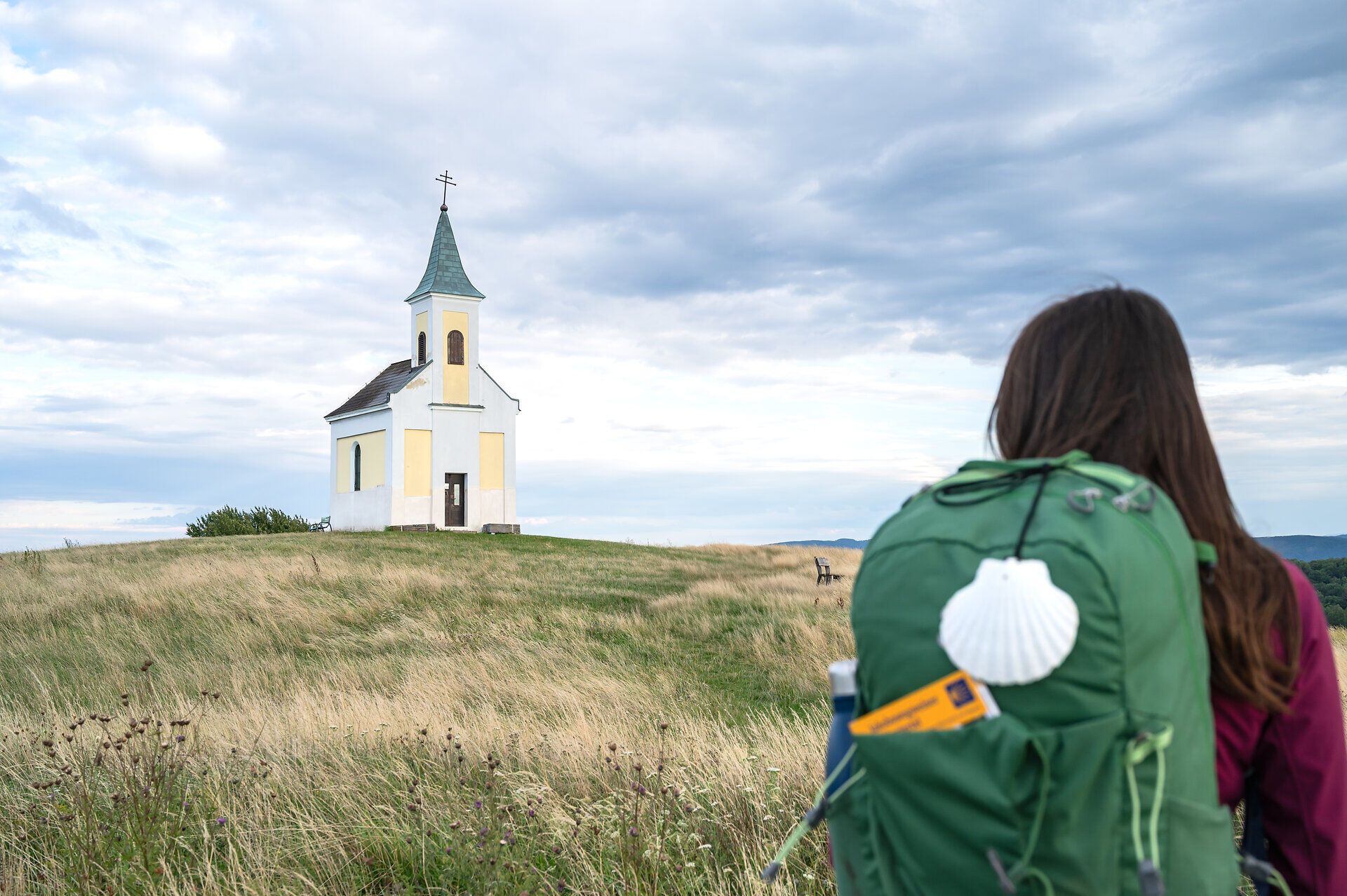 Eine Pilgerwanderin trägt einen Rucksack mit Jakobsmuschel, Reisehandbuch und Trinkflasche und blickt zur Kapelle am Michelberg.