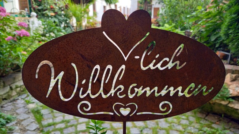Metallschild mit der Aufschrift 'Herzlich Willkommen' in einem Garten.