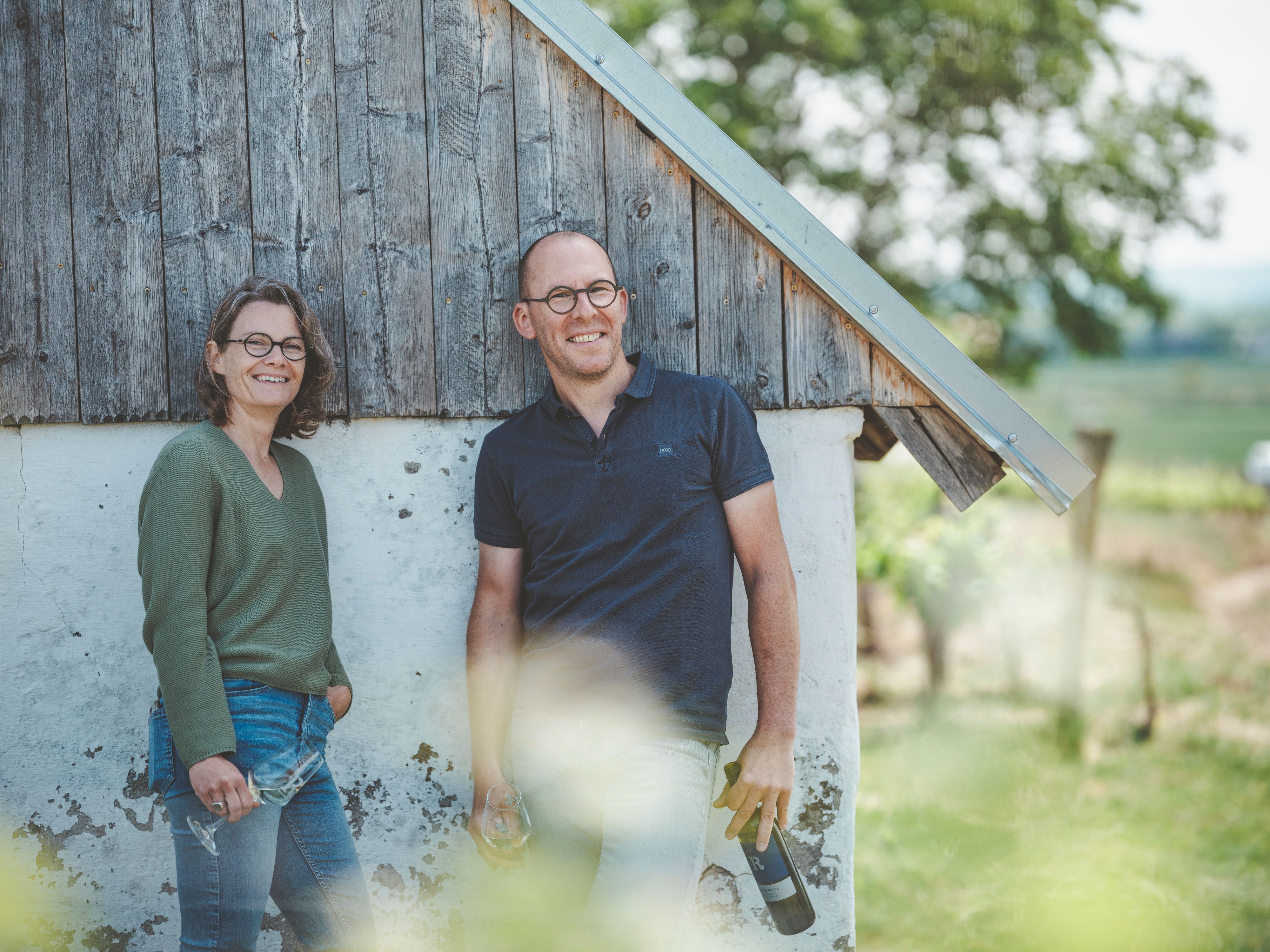 Two people stand smiling in front of a wooden wall outside.