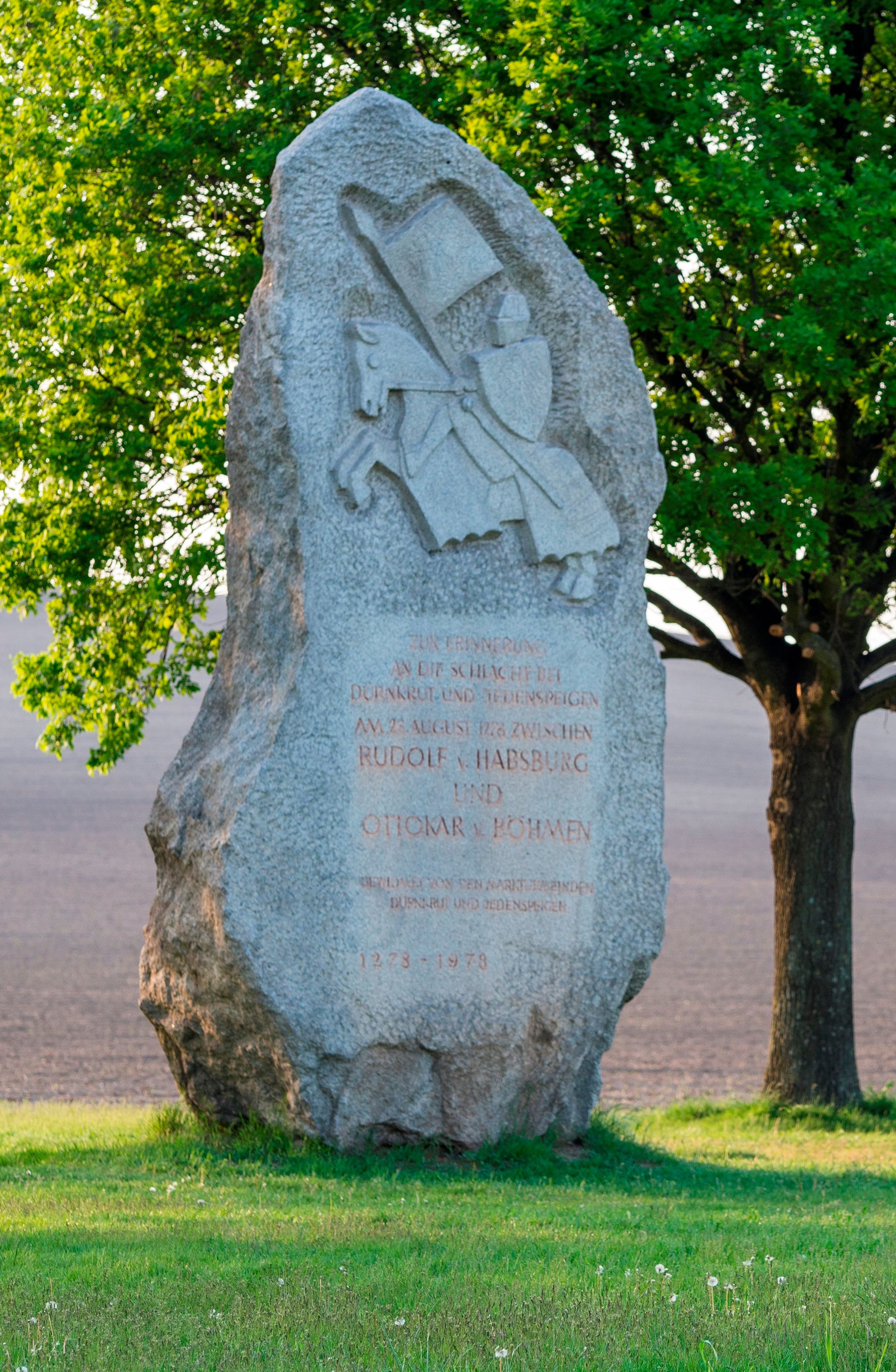 Gedenkstein mit Ritterrelief und Inschrift, umgeben von Bäumen und Wiese.