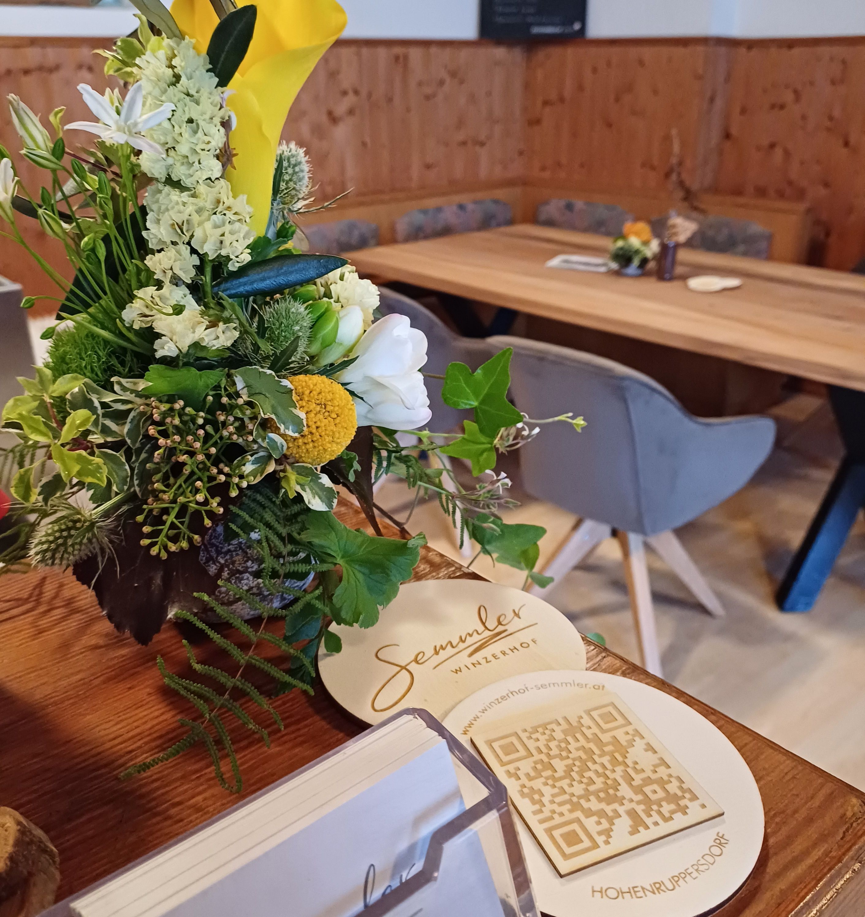 Ein Blumenarrangement auf einem Tisch im Winzerhof Semmler mit QR-Code und Visitenkarten.