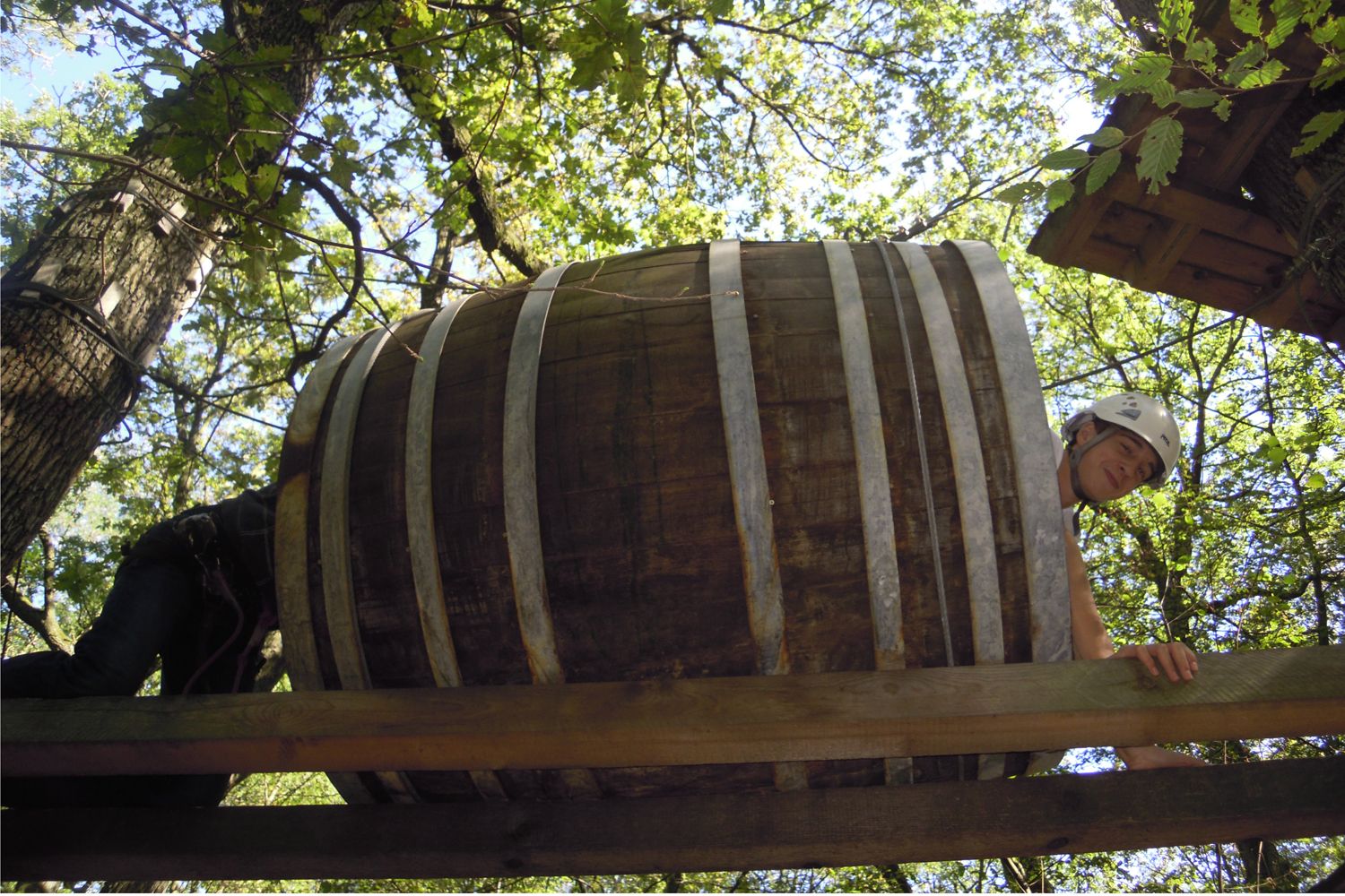 Person mit Helm kriecht durch ein großes Holzfass in einem Kletterpark.