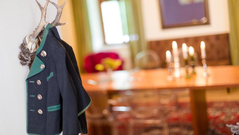 Ein traditioneller Janker hängt an einem Geweih an der Wand. Im Hintergrund ein Tisch mit Kerzenleuchtern und ein Fenster.