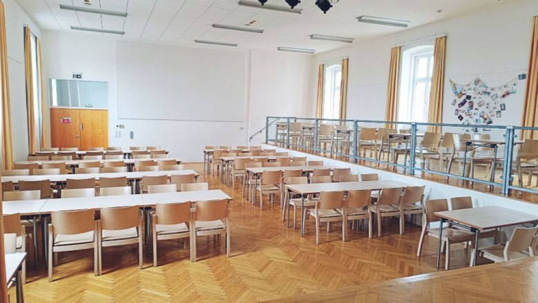Ein leerer Saal mit Holzboden, Tischen und Stühlen, einer Bühne und Beleuchtung an der Decke.