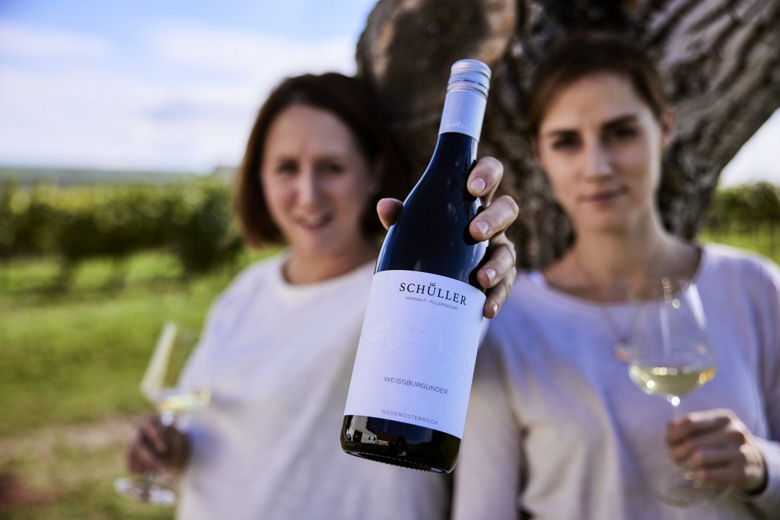 Zwei Personen halten eine Flasche Schüller Weißburgunder und Weingläser vor einem Baum in einem Weinberg.