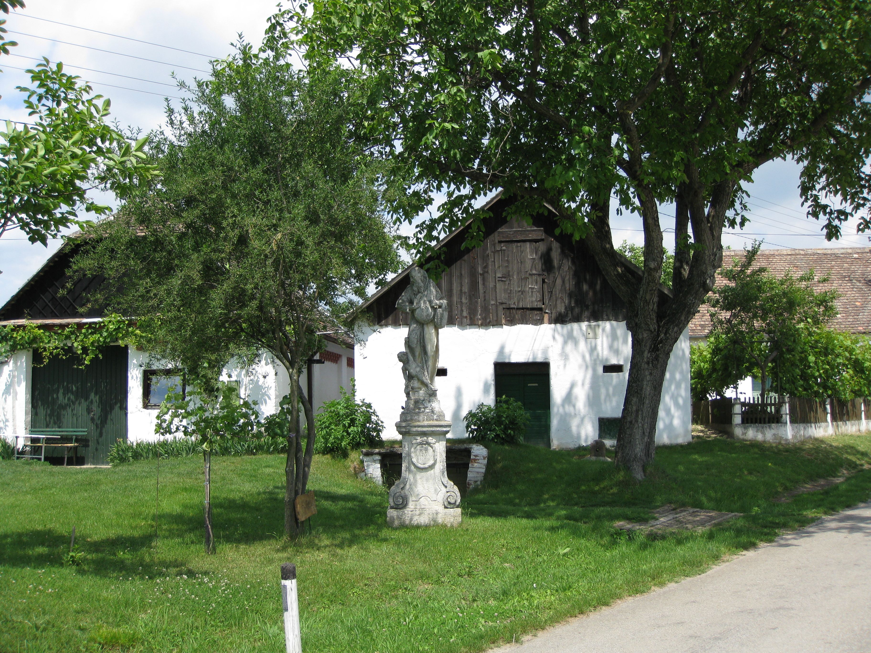Ein weißes Gebäude mit Holztor und Statue im Vordergrund, umgeben von Bäumen und grünem Rasen.