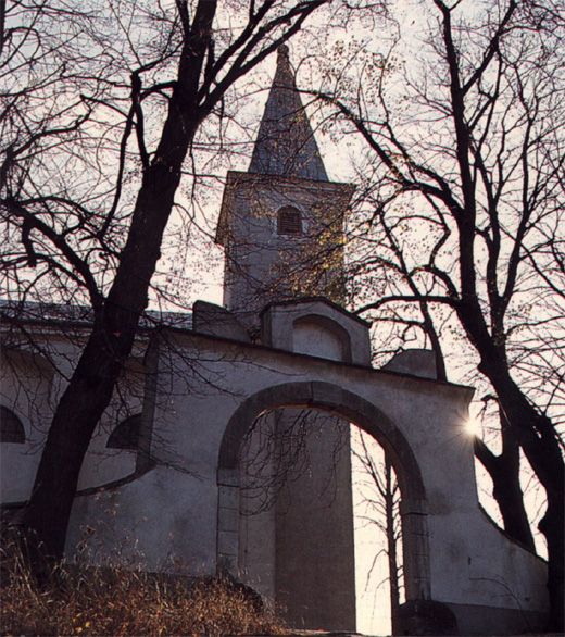 Kirche mit Turm und Bogen, umgeben von kahlen Bäumen im Gegenlicht.