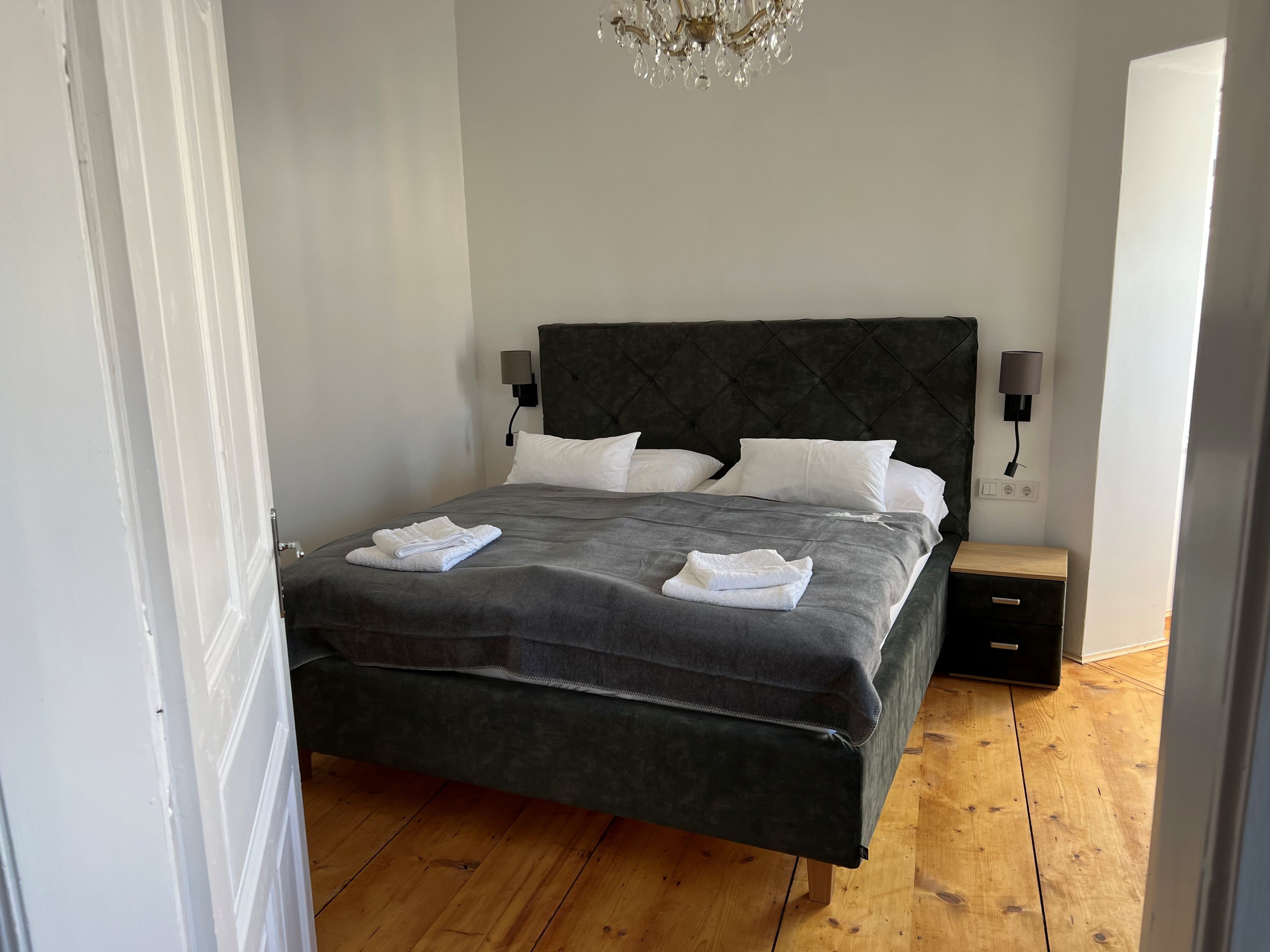 Ein gemütliches Schlafzimmer mit einem großen Bett, grauer Bettwäsche und weißen Handtüchern. Ein Kronleuchter hängt von der Decke.