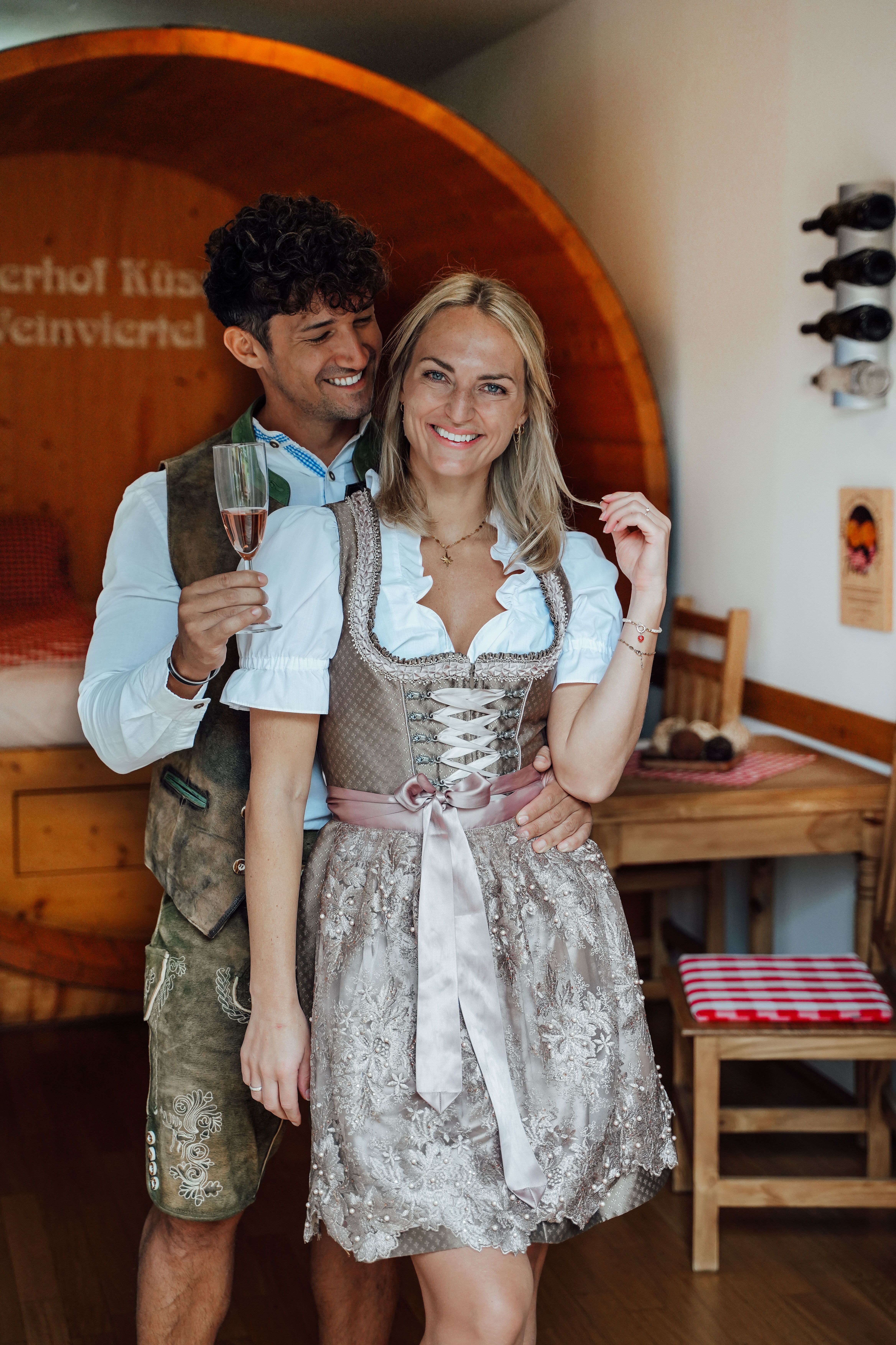 Paar in Tracht im Winzerhof Küssler: Der Mann hält ein Glas Rosé, die Frau lächelt in die Kamera. Im Hintergrund Weinfass und Holzmöbel.