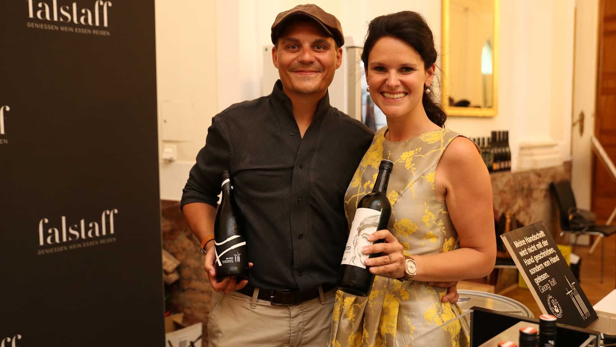 Two people holding bottles of wine in front of a Falstaff poster.