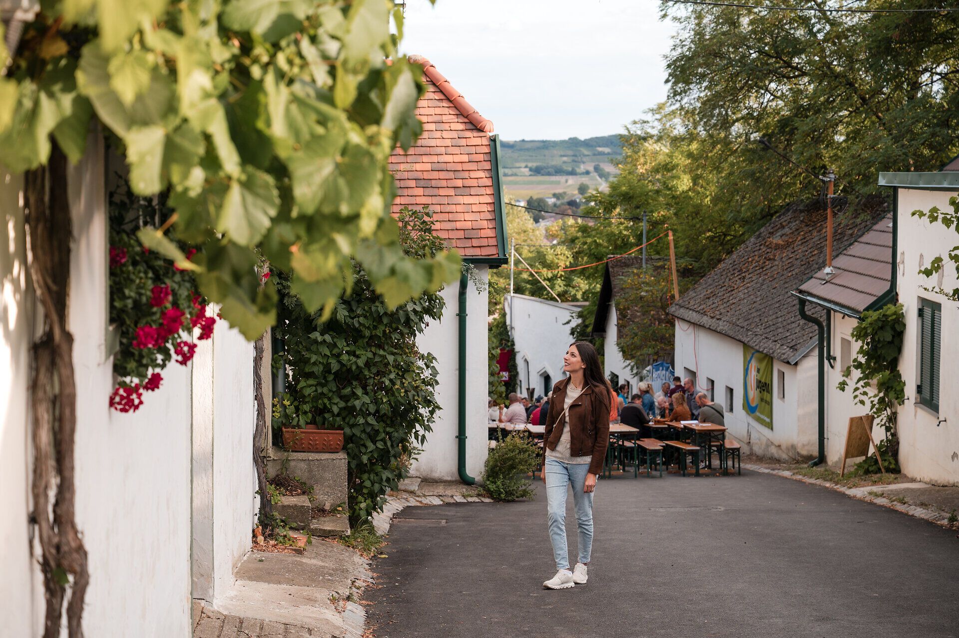 Eine Frau spaziert durch eine Kellergasse und blickt sich um, während im Hintergrund geselliges Treiben bei einem Kellergassenfest herrscht.