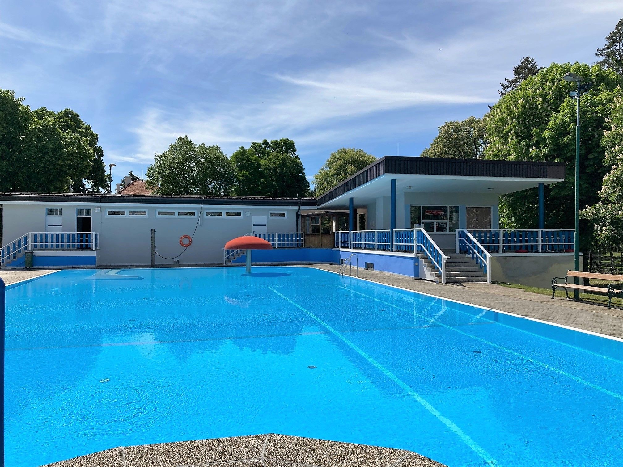 Ein Freibad mit klarem Wasser, umgeben von Bäumen und einem Gebäude mit blauen Geländern.