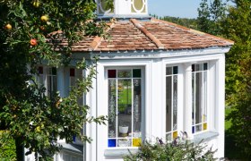Ein wei&szlig;er Pavillon mit bunten Fenstern in einem Garten, umgeben von B&auml;umen und einem Zaun.
