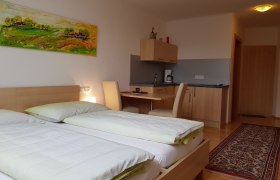 Ein modernes Zimmer mit zwei Betten, einem Schreibtisch, einer kleinen K&uuml;chenzeile und einem bunten Gem&auml;lde an der Wand.