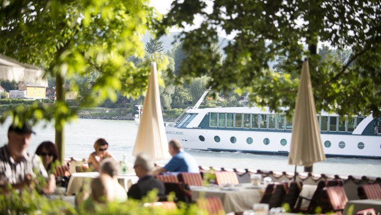 Ein Flusskreuzfahrtschiff fährt auf der Donau, im Vordergrund sitzen Menschen in einem Café unter Bäumen.