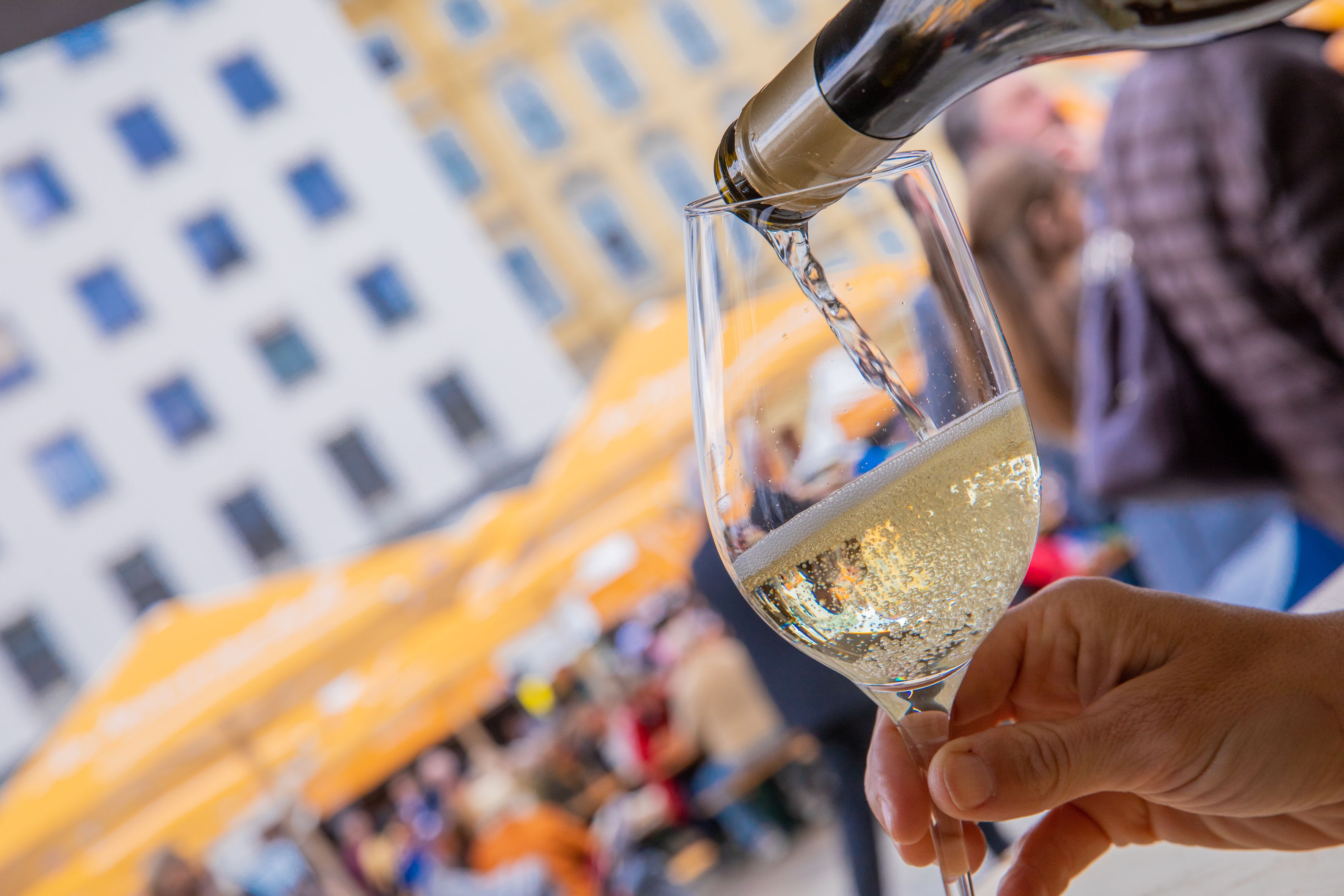Im Vordergrund wird gerade ein Glas Weinviertler Sekt eingeschenkt, im Hintergrund sieht man Personen, welche Weinviertler Getränke und Kulinarik in einer Innenstadt genießen. 