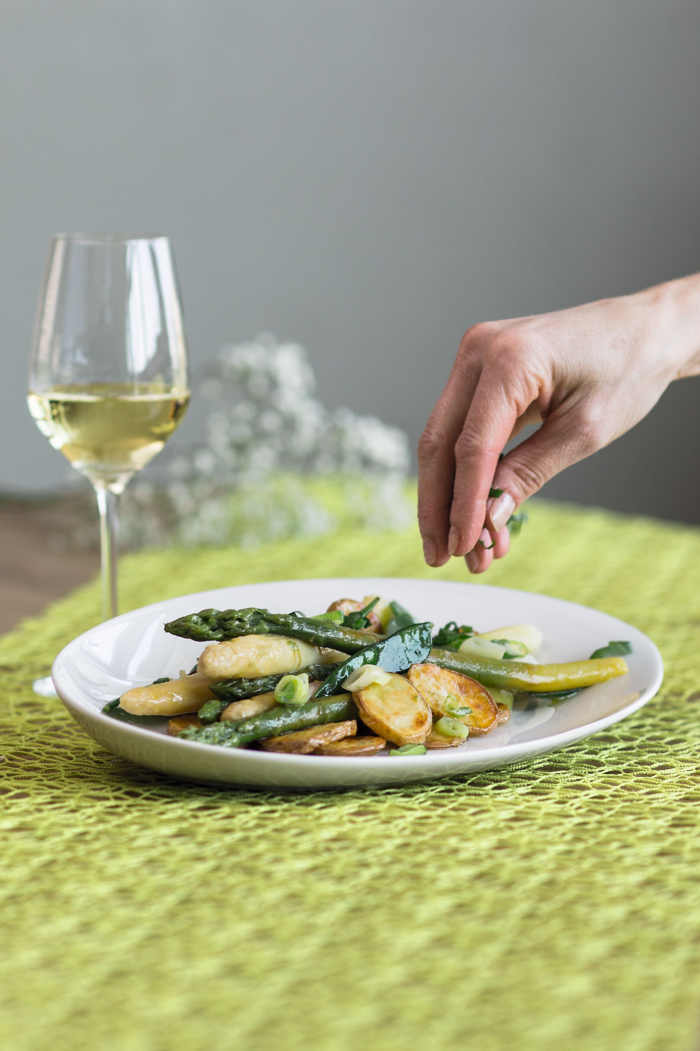 Ein Teller mit Spargel und Erdäpfeln steht neben einem Glas Weißwein auf einem Tisch. Eine Hand streut Kräuter darüber.