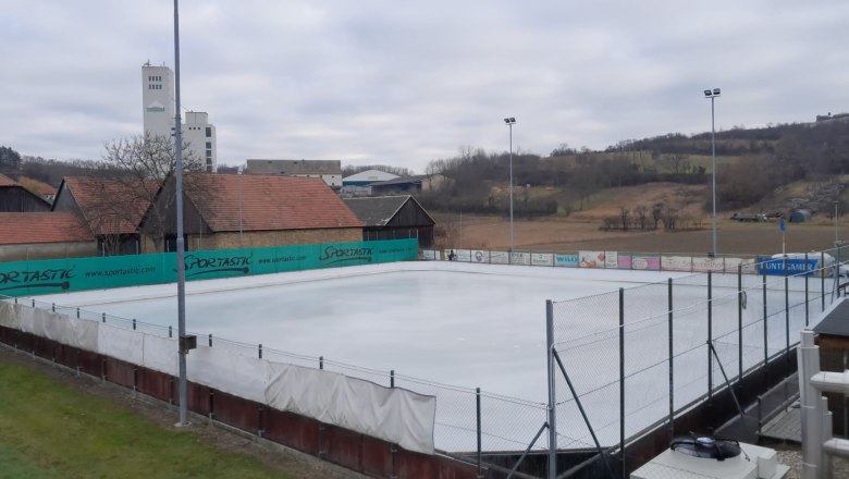 Ein leerer Eislaufplatz mit umliegenden Gebäuden und Hügeln im Hintergrund.