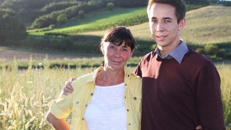 A woman and a young man stand in a rural landscape and smile at the camera.
