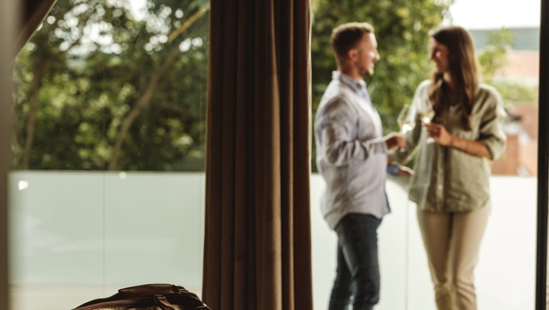 Ein Paar steht auf einem Balkon und h&auml;lt Weingl&auml;ser, im Vordergrund liegt eine Reisetasche auf einem Bett.