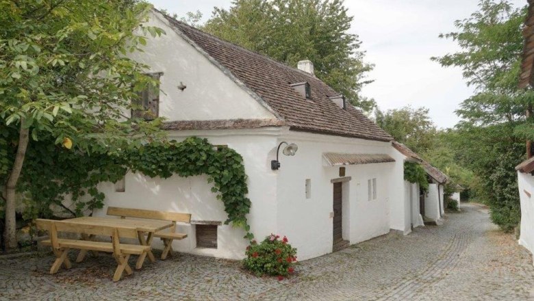 Weißes Kellerstöckl mit Weinstöcken und Holztisch auf Kopfsteinpflasterstraße.