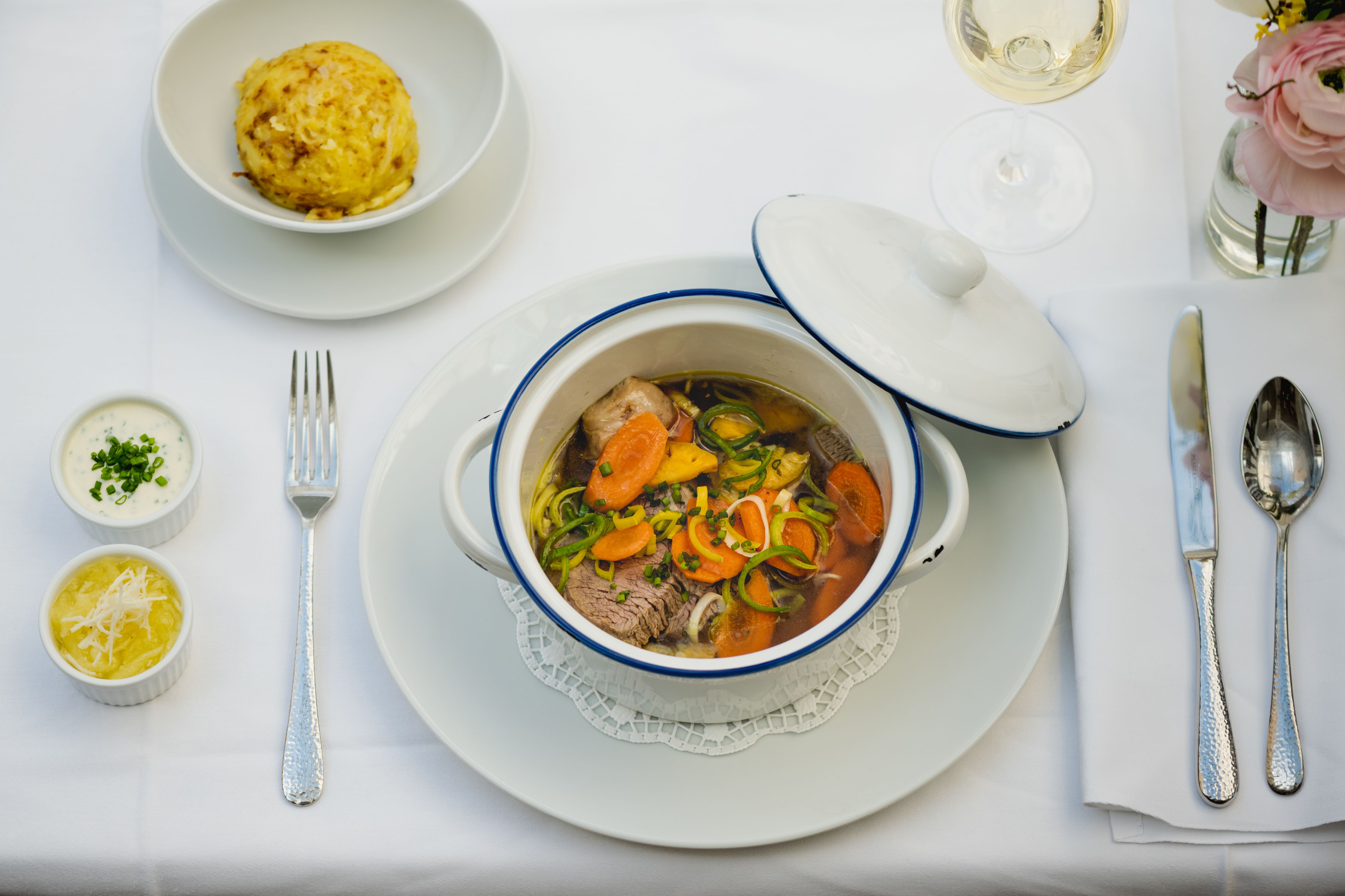 Ein gedeckter Tisch mit einem Eintopf in einem weißen Topf, einem Knödel, zwei Saucen und einem Glas Weißwein.