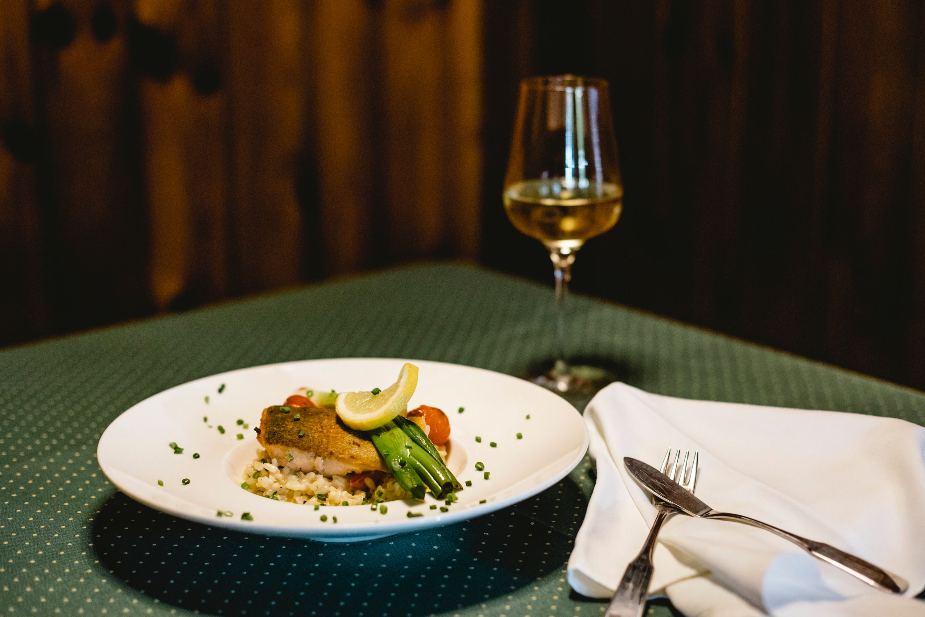 Ein Teller mit Lachsforelle und Risotto, garniert mit Zitrone und Gemüse, steht auf einem gedeckten Tisch neben einem Glas Weißwein.