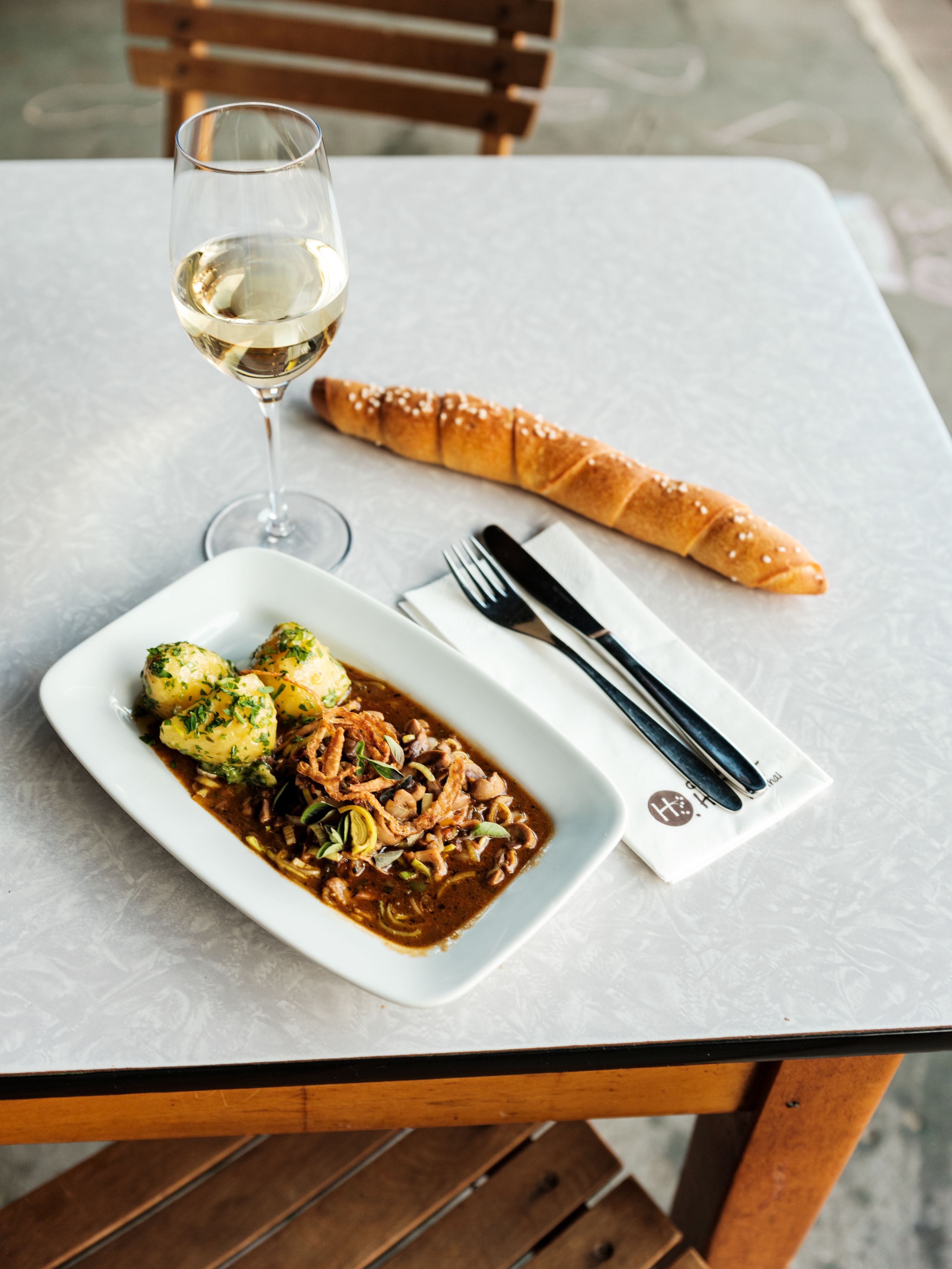 A table with a glass of white wine, a plate with a meat dish and dumplings, a long bread roll and cutlery on a napkin.