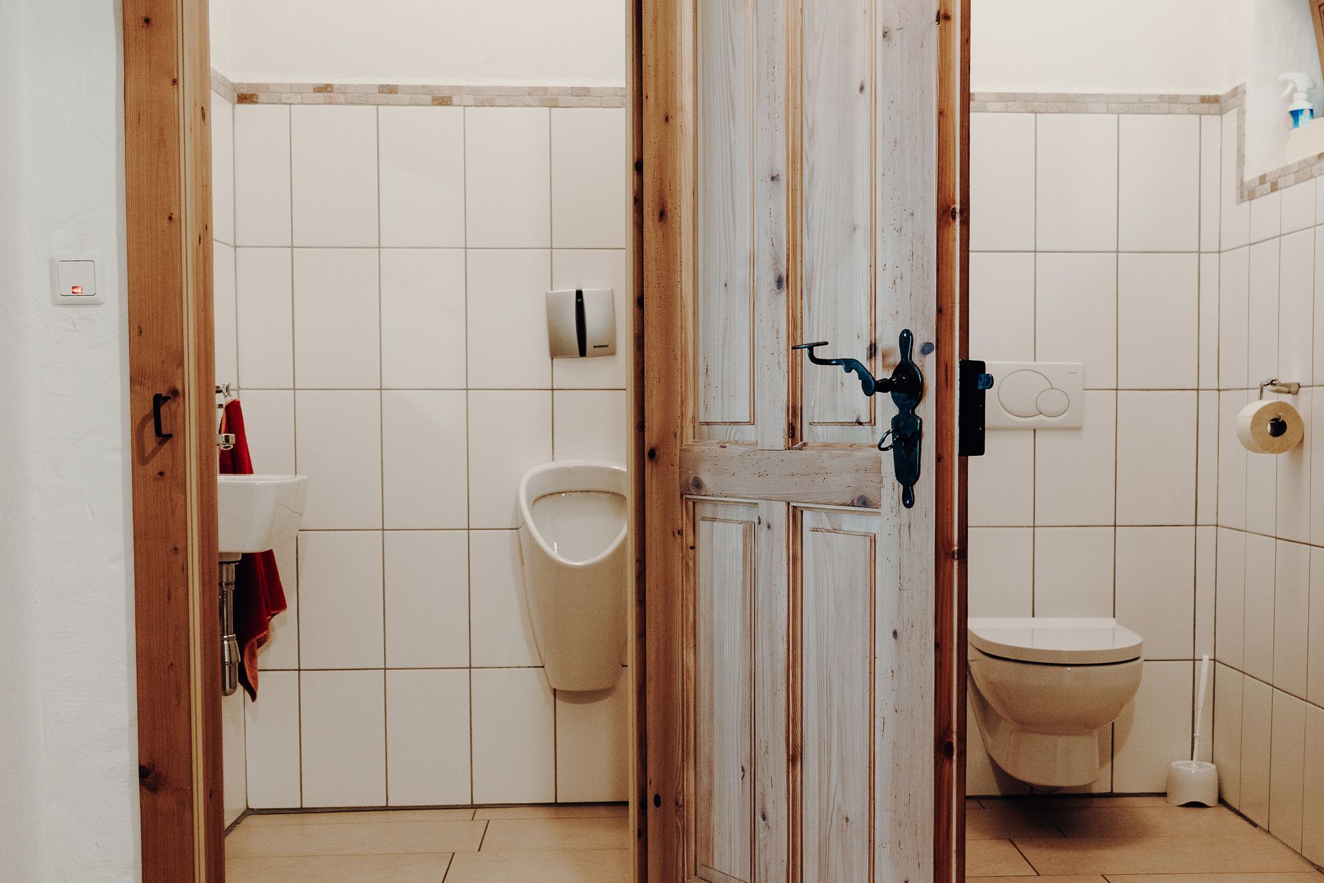 Zwei Toilettenräume mit Holztür, links ein Urinal und Waschbecken, rechts eine Toilette.