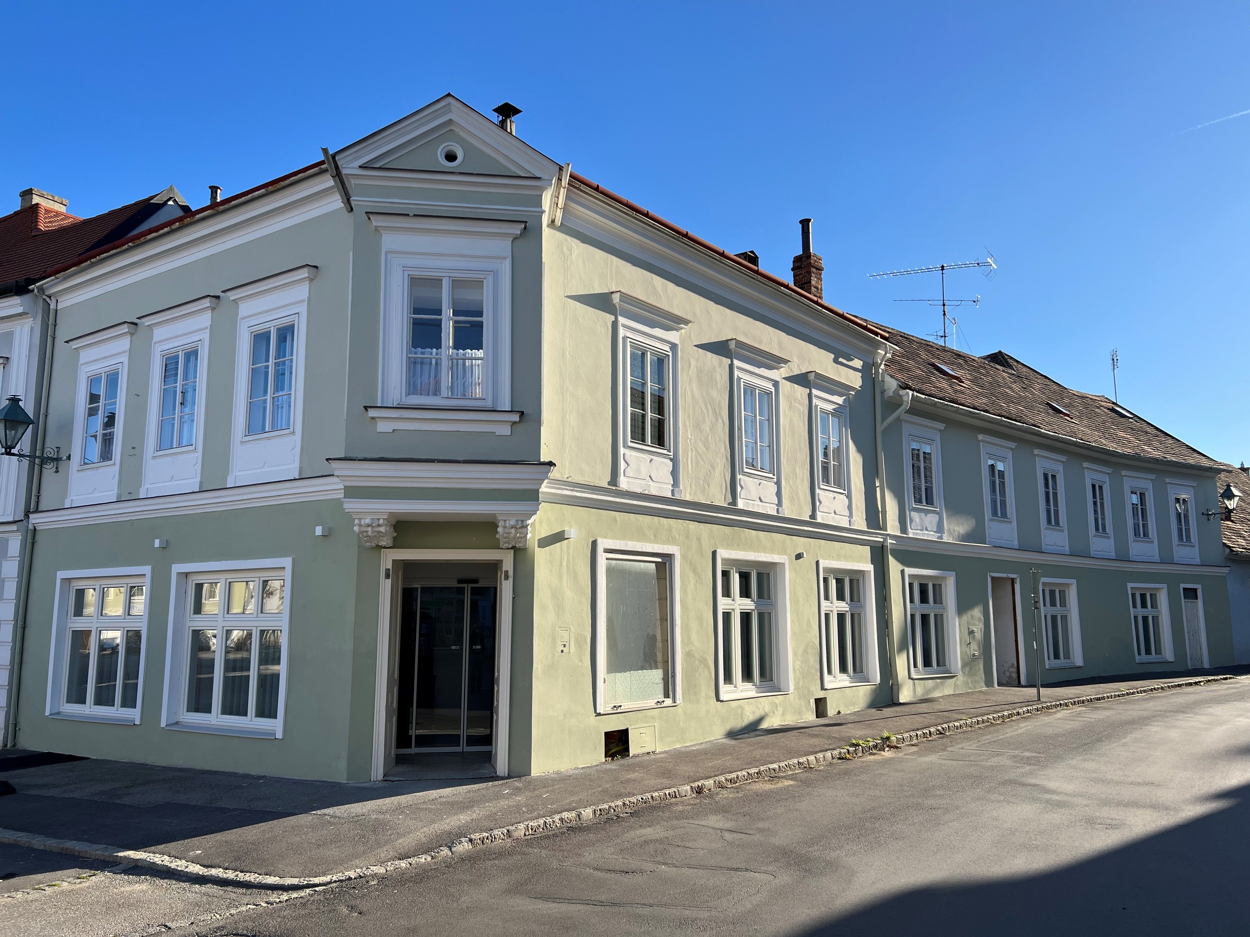 Ein zweistöckiges, pastellgrünes Gebäude mit weißen Fensterrahmen an einer Straßenecke bei klarem Himmel.