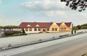 Außenansicht eines Weinguts mit gelbem Gebäude und rotem Dach, umgeben von Weinreben und einer Straße im Vordergrund.