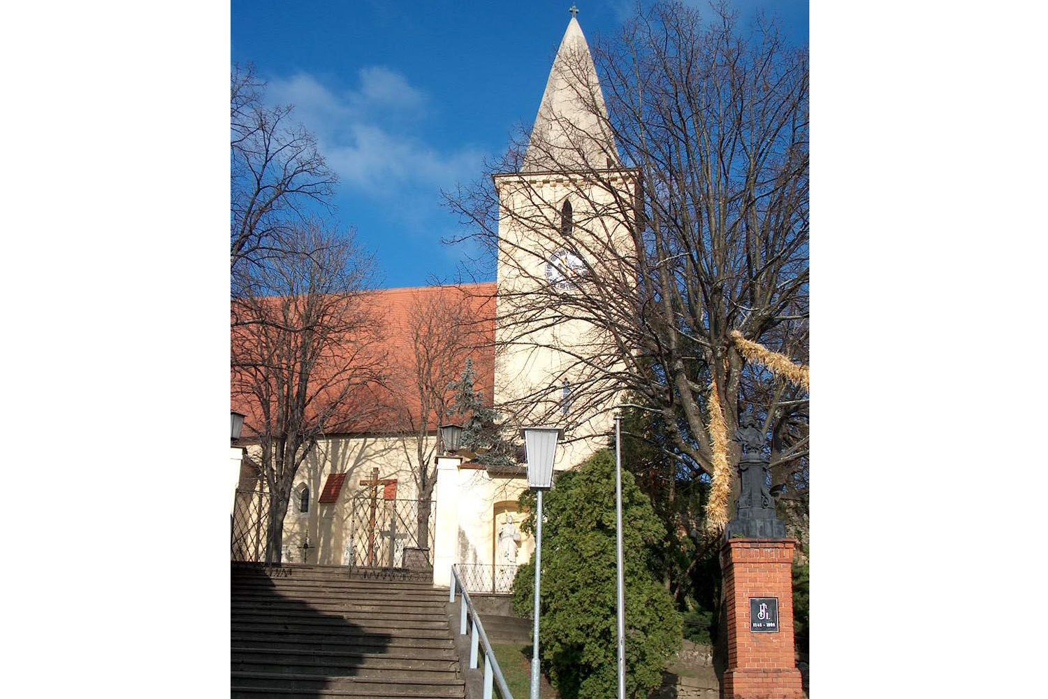 Pfarrkirche mit Turm und roten Dachziegeln, umgeben von Bäumen und Treppen im Vordergrund.