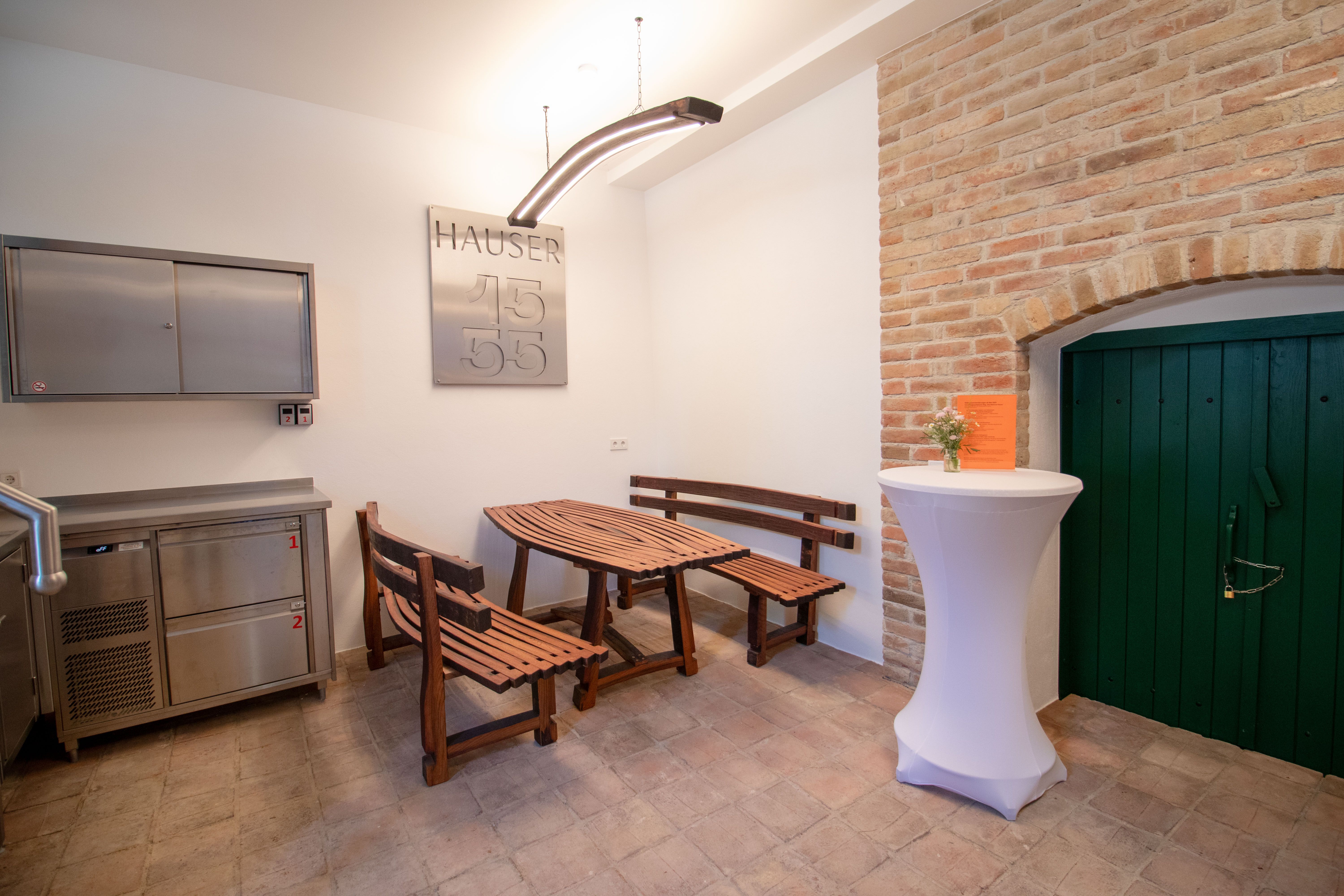 A rustic room with wooden benches, a wooden table, a bar table with flowers and a green door in a brick wall.