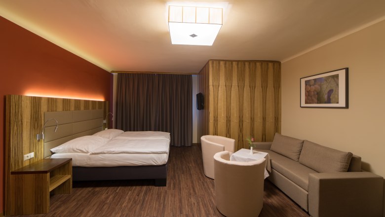 Modern eingerichtetes Hotelzimmer mit Doppelbett, Sofa, zwei Sesseln und einem kleinen Tisch. Holzboden und warme Beleuchtung.