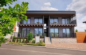 Moderne zweistöckige Apartmentanlage mit Balkonen und Treppenaufgang, umgeben von Pflanzen und Bäumen.