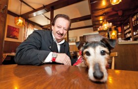 Ein Mann in einem Gasthaus lächelt, während ein Hund mit rotem Halstuch auf dem Tisch liegt.
