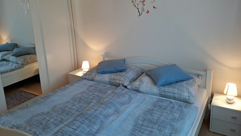 Gemütliches Schlafzimmer mit Doppelbett, blauer Bettwäsche, Nachttischen mit Lampen und einem Wandtattoo mit Blumen und Vögeln.