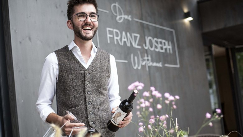 Mann mit Brille und Weste h&auml;lt Weinflasche vor Restaurant 'Franz Joseph'.
