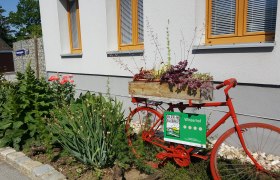 Ein rotes Fahrrad mit Blumenkasten steht vor einem Gebäude mit Garten.
