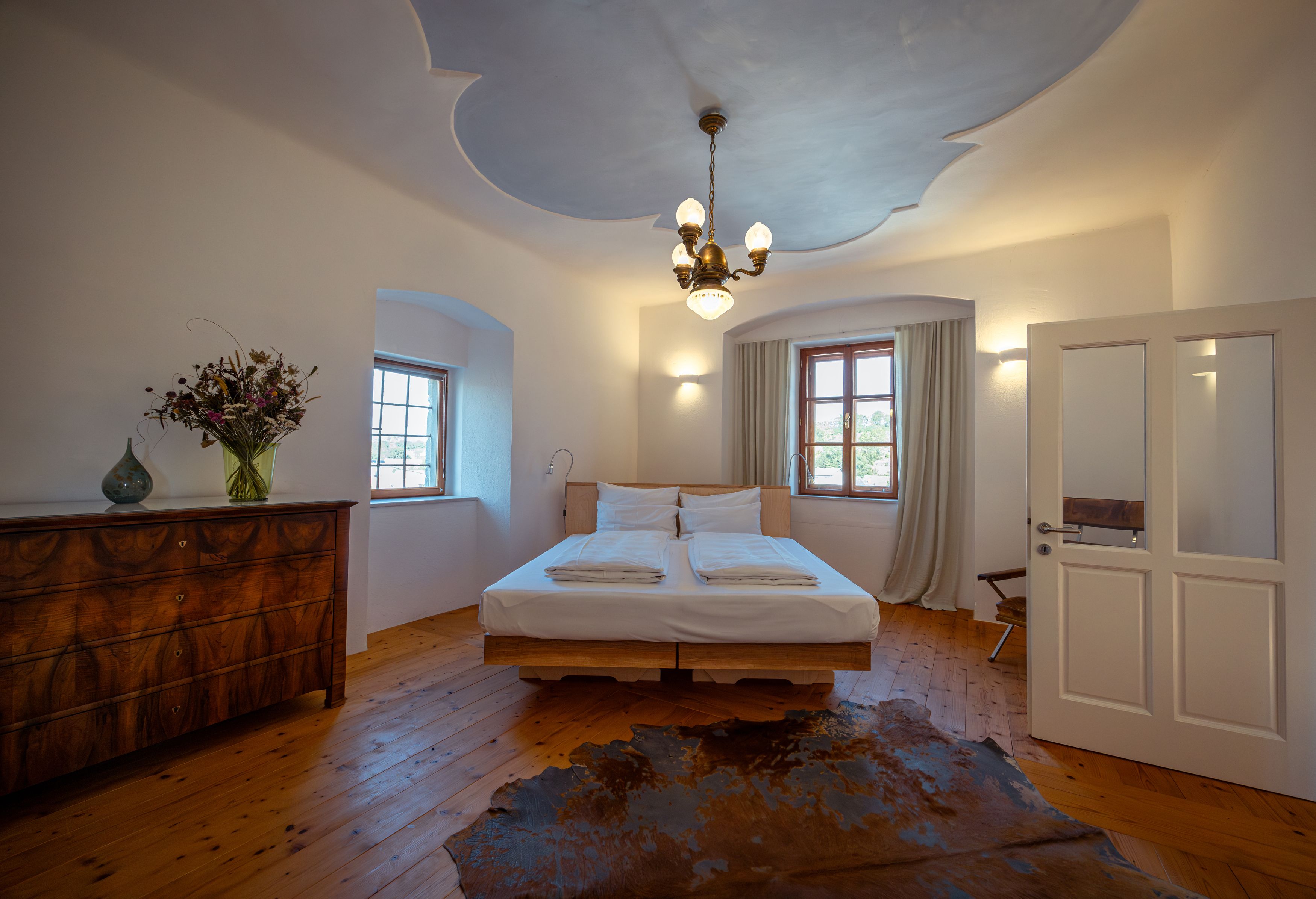 Ein gemütliches Schlafzimmer mit Holzboden, Doppelbett, antiker Kommode und Blumenstrauß. Deckenleuchte und Fenster mit Vorhängen.