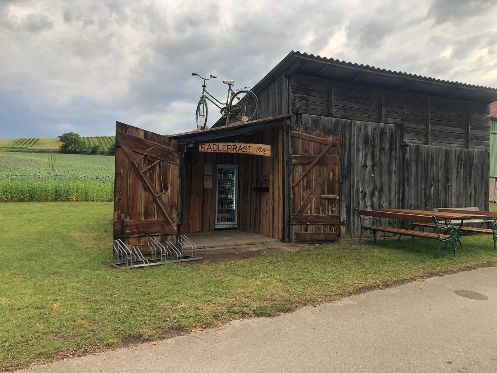 Eine rustikale Holzhütte mit der Aufschrift 'Radlerrast' und einem Fahrrad auf dem Dach, umgeben von Feldern und einer Bank.