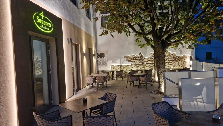 Außenterrasse eines Restaurants mit Tischen und Stühlen, beleuchtet bei Dämmerung, Baum im Hintergrund.