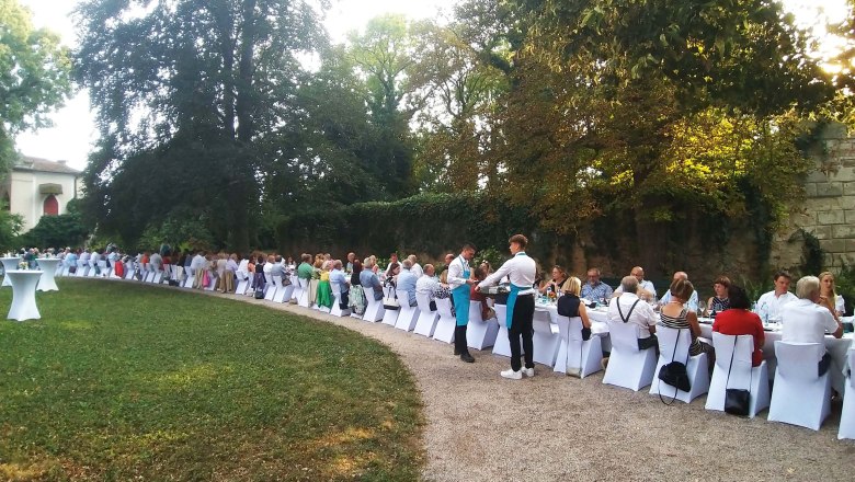 Langes Bankett im Freien mit vielen Gästen an weiß gedeckten Tischen, umgeben von Bäumen.