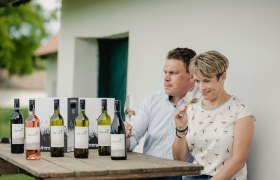 Zwei Personen sitzen an einem Tisch mit Weinflaschen und probieren Wein.