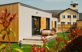 Moderne Appartements mit Holzverkleidung und Weinfässern auf der Terrasse, umgeben von grünem Rasen und einem Zaun.