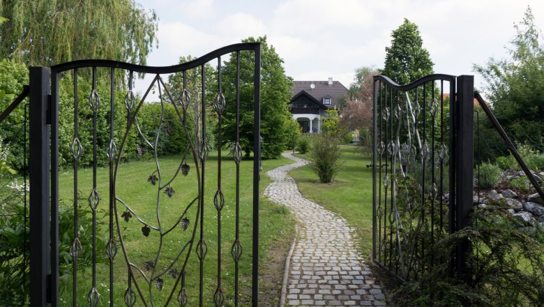 Ein offenes schmiedeeisernes Tor führt zu einem gepflasterten Weg, der durch einen grünen Garten zu einem Haus im Hintergrund führt.