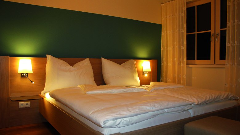 Ein gem&uuml;tliches G&auml;stezimmer mit einem Doppelbett, zwei Nachttischlampen und einem Fenster mit Vorh&auml;ngen.