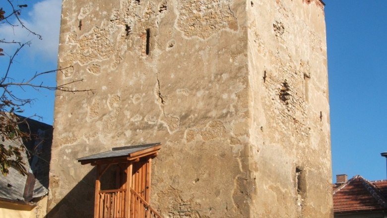 Ein alter Wehrturm mit hölzerner Treppe und Geländer, umgeben von Gebäuden, unter blauem Himmel.